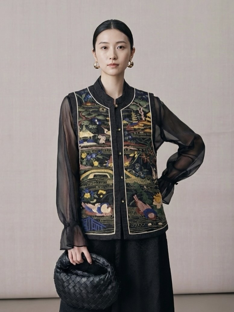 Woman wearing a black embroidered vest over a sheer black top, holding a black handbag against a plain background.