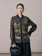 Woman wearing a black embroidered vest over a sheer black top, holding a black handbag against a plain background.